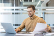 © Liubomir - Productivity and corporate workflow, smiling businessman working on laptop with documents in modern office.