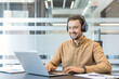 © Liubomir - Remote work and digital communication, smiling man using laptop and headphones in modern office. Professional employee typing on computer and attending video conference, freelancer collaborating.