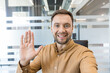 © Liubomir - Remote work and virtual meeting concept, smiling businessman waving hand during video call in modern office. Professional communication and digital technology in corporate culture.
