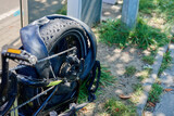 A bicycle is parked on a sidewalk with a flat rear tire. Grass grows nearby under clear skies. The setting is typical for an urban area