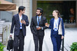 © ultramansk - A multiethnic business team of three colleagues walk past luxury shopfronts holding takeaway coffee outside a glass office tower in a modern Asian financial district
