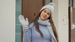 © Krakenimages.com - Woman brunette smiling waving gloved hand, wearing grey knit hat, scarf and sweater, standing by a building entrance door; friendly greeting.