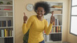 © Krakenimages.com - Woman in yellow sweater smiling and giving thumbs up beside tall bookshelves and a wall clock in a home study inside a building; joy confidence.