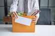 © Charlie's - Businessman Sending Resignation Letter to Boss While Holding Cardboard Box Beside Office Desk After Career Change or Job Loss.