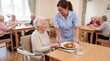 © Marina Shvedak - Woman nurse serving lunch to happy senior woman in care home dining room. Elderly residents eating meal together at retirement facility. Healthcare and elderly care concept.