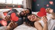 © Eve - Playful interracial couple lying in bed holding big red paper hearts and laughing with heart bokeh background, ideal commercial banner for Valentine's Day romantic getaway promotions.