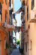 © dsbedarieva - Laundry hangs on clotheslines stretched across a narrow alley in Kerkira, Greece. Tall, colorful buildings line the historic Mediterranean street.