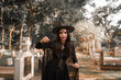 © Kieng - A mysterious witch in a black costume and pointed hat stands in a dark cemetery, surrounded by tombstones and deep shadows, creating an eerie Halloween atmosphere.