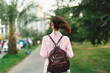 © SHOTPRIME STUDIO - Young woman with striped shirt walks away through a green park carrying a brown backpack, capturing a simple moment of casual exploration.