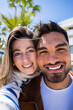 © CarlosBarquero - Vertical. Close portrait of a happy couple smiling directly at camera while standing outdoors in a bright modern residential area with palm trees and blue sky.