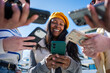 © CarlosBarquero - Outdoor lifestyle scene of a Black woman using a smartphone while surrounding hands hold phones and money, suggesting online payment and social finance.