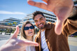 © CarlosBarquero - Young man and woman smile at camera while making a hand frame outdoors in a bright residential district with modern architecture and clear sky.