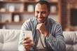 © Prostock-studio - Cheerful Black Man Smiling While Using Smartphone, Sitting Comfortably On Couch At Home. Engaging With Phone In Relaxed Setting, Enjoying Leisure Time And Connected Experience.