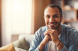 © Prostock-studio - Smiling Young Black Man Relaxing At Home, Sitting On Sofa With Hands Under Chin, Displaying Happiness And Contentment In Cozy Living Room Setting, Signifying Warm Connections And Lifestyle.