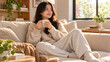 © Somadi - Happy woman in cozy knitwear drinking coffee or tea while sitting on a comfortable couch in a sunlit room