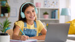 © Vyatcheslav - Happy young girl with headphones studying online with a laptop. Smiling child writing in a notebook at her desk. E-learning and homeschooling concept