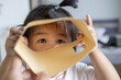 © banjongseal324 - Cute Asian Toddler Girl Peering Through Open Brown Cardboard Sleeve Frame,A close-up lifestyle photograph of a playful cute young Asian toddler girl peering through an open brown paper envelope.