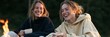 © Andrii Zastrozhnov - Two young women laughing by a campfire holding mugs outdoors at dusk enjoying a cozy moment for lifestyle social media header