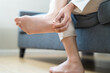 © KMPZZZ - Foot pain concept, close up hand of young woman rubbing, massaging sore feet area of heel, girl suffering on sofa, couch at home. Discomfort painful feet ache from walking for long. Physical injury.