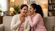 © Omni - Indian daughter whispering a secret into her laughing mother's ear while sitting on a couch at home