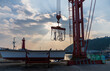 © Travel 'n' Lifestyle - View of a red industrial crane lifting equipment over a boat on a trailer at a harbor, with a red lighthouse in the background under a cloudy sky in Busan, Busan, South Korea.