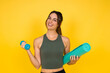 © Daniel - Young woman smiling and feeling confident while holding a light blue dumbbell and a rolled-up teal yoga mat, representing a healthy and active lifestyle on a yellow background
