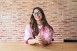 © Daniel - Young confident woman wearing glasses and a pink shirt. Smiling and looking directly at the camera. Sitting at a wooden table in front of a brick wall. Representing professionalism and approachability