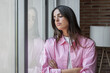 © Daniel - A young woman in a pink striped shirt stands at a window with arms crossed, gazing outside in a pensive, sad mood. The indoor scene conveys loneliness, reflection and quiet contemplation.