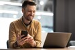 © Prostock-studio - Smiling Caucasian Male In Business Casual Attire Engaged With Smartphone While Using Laptop At Modern Office Desk, Showing Professionalism And Productivity In Work Environment.