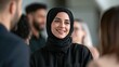 © 69 - Young woman wearing black hijab smiling and interacting happily with diverse group in professional casual social gathering indoors