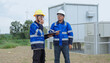 © Father_Studio - Wind energy technician reports by handheld radio while an engineer holds inspection documents, coordinating field observations beside utility equipment during a renewable power site check.