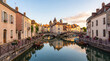 © Vivid Pixel - Charming historic canal town at sunset with stone bridges, moored wooden boats, pastel houses, flower boxes and calm water reflections under a soft evening sky
