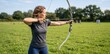 © ProNoJoy - Woman in a grey t-shirt aiming a recurve bow at a target in a grassy field, outdoor archery practice session on a sunny day with trees in the background