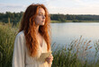 © yollieee - Thoughtful young red-haired woman holding a letter by a quiet lake at sunset, creating a nostalgic concept of memories, longing and personal communication.