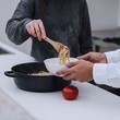 © AI-Universe - Hände servieren dampfende Spaghetti aus schwarzer Pfanne
