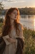 © yollieee - Thoughtful young red-haired woman holding a letter by a quiet lake at sunset, creating a nostalgic concept of memories, longing and personal communication.