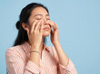 © JoelBourgoin - Generative AI studio portrait photography of a young Asian woman wearing a striped shirt rubbing her tired eyes with her hands against a solid light blue background.