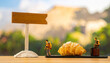 © SolaruS - Miniature figurines of travelers with backpacks stand next to a golden croissant and a wooden signpost. This whimsical, no people travel concept evokes a sense of adventure and culinary tourism.