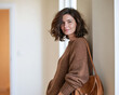 © Anna - A stylish brunette woman poses casually in an oversized brown knit sweater with a warm-toned shoulder bag, leaning against a wall in a bright minimalist interior.