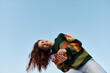 © SHOTPRIME STUDIO - Young woman in a colorful striped sweater smiles while taking a selfie with her phone against a blue sky. Wind blows her long hair. Concept of joy, communication, and mobile lifestyle.