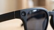© Sobia - Macro close-up of matte black smart glasses frame with integrated camera lens in natural daylight on neutral background