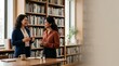 © Alva - Two professional women having a friendly conversation in a modern library office setting.