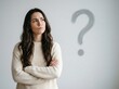 © Pixels Stock - Confused young woman in beige knitted sweater stands with arms crossed looking towards hand painted grey question mark on wall as she ponders a difficult decision.