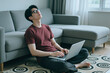 © DragonImages - Asian young adult man sitting on floor with laptop, leaning back against sofa with eyes closed, feeling tired. Image represents remote work burnout, suitable for mental health campaigns