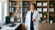 © Aurora - Confident female doctor in white coat smiling, holding stethoscope in her office with bookshelves and diplomas in background