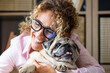 © simona - Woman with curly hair and glasses hugging a happy senior pug, smiling warmly while cuddling her pet dog, conveying affection, comfort, companionship and a joyful domestic moment.