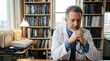 © Gogo - Thoughtful senior doctor in white coat sitting at desk in office, hands clasped with stethoscope, surrounded by bookshelves filled with medical books.