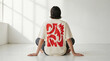© kinkin - Back view of a person sitting on the floor wearing a white t-shirt with a red abstract graphic design in a bright minimalist room