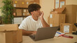 © Krakenimages.com - Man touches face while checking laptop and packages in a small business building; entrepreneurial planning contemplation.