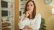 © Krakenimages.com - Woman touching chin with crossed arms in building office interior with bookshelf, clock and plant visible; contemplation.